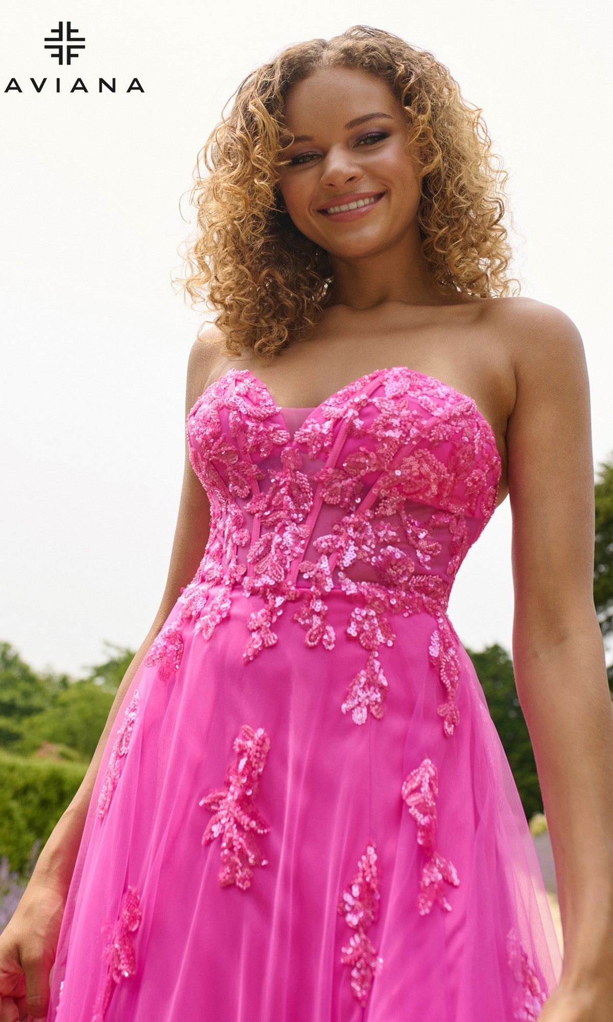 A woman in the Faviana 11474 long prom dress, featuring sequin, lace, and beaded appliqué details, stands smiling outdoors. The Faviana logo is visible in the top left corner.
